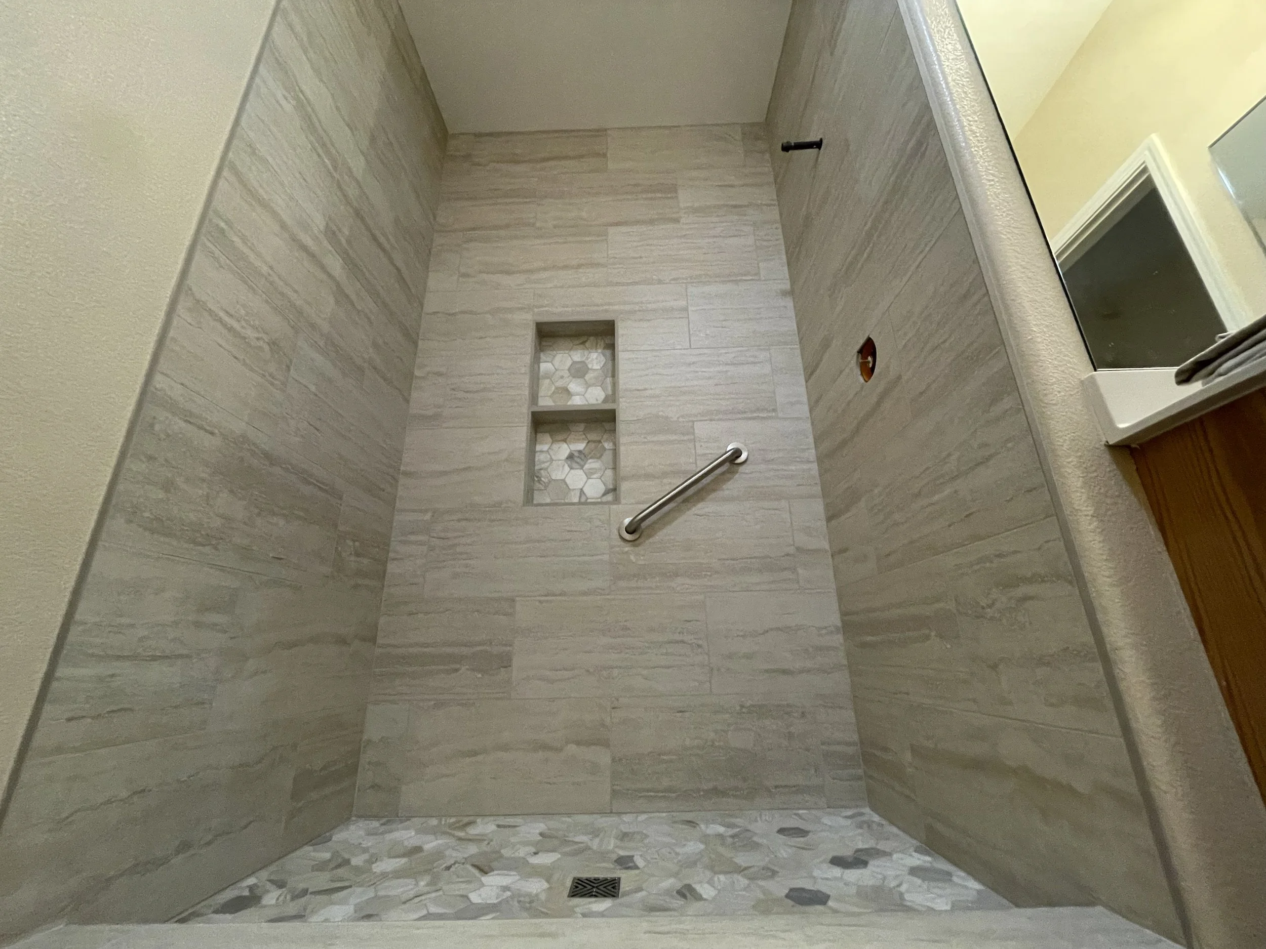Indoor shower with beige tile walls, small built-in shelves with hexagon tiles, a metal grab bar, and a drain on the tiled floor.