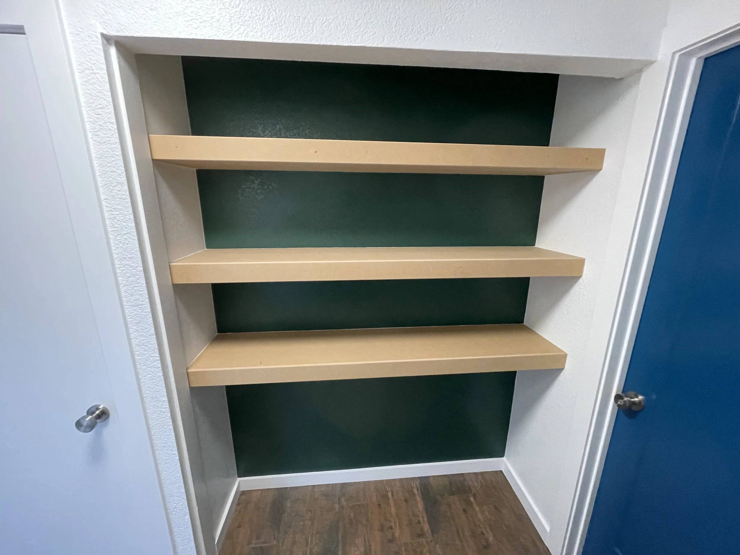 Empty custom built-in shelf with three wooden shelves against a expertly painted green wall, white walls on either side, and a brown tile wood floor.