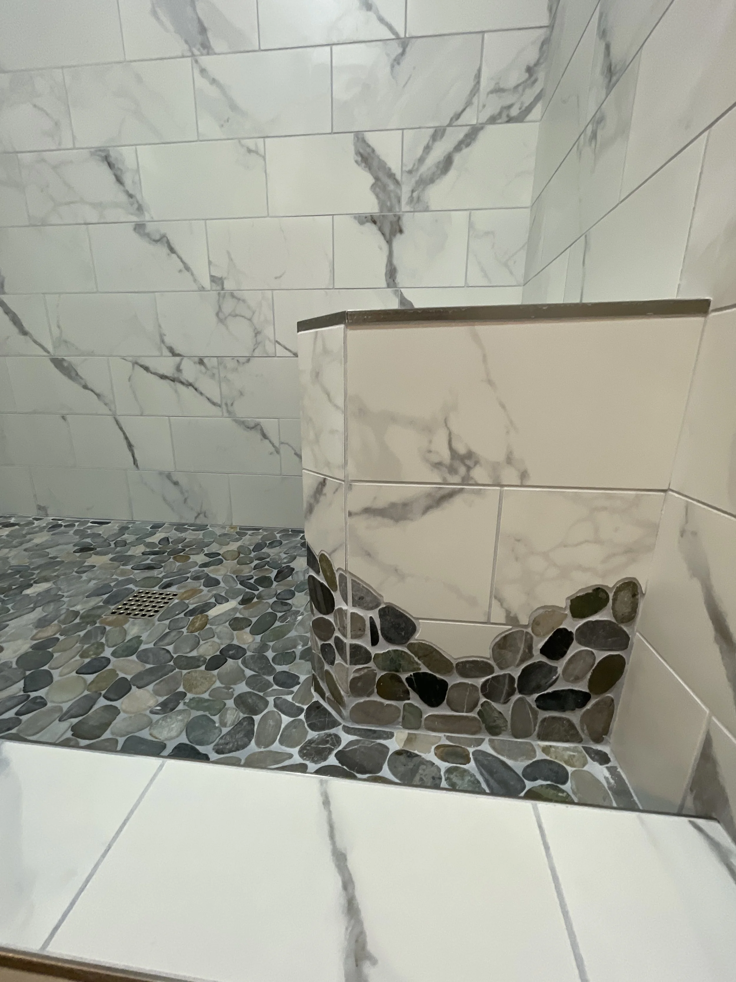 A shower corner with white and gray marble tiles on the walls and a pebble mosaic floor, featuring a drainage grate.