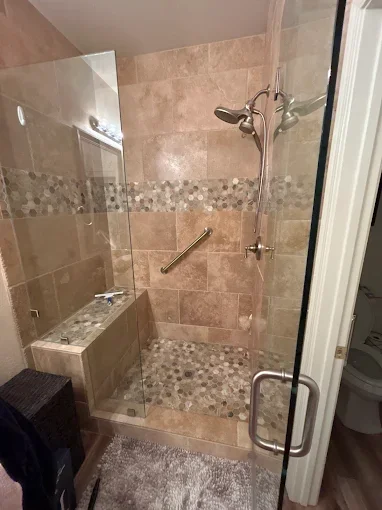 Bathroom with a walk-in shower enclosed by glass, featuring beige and pebble tile accents, a handheld showerhead, a built-in bench, and a toilet visible in an adjacent room.
