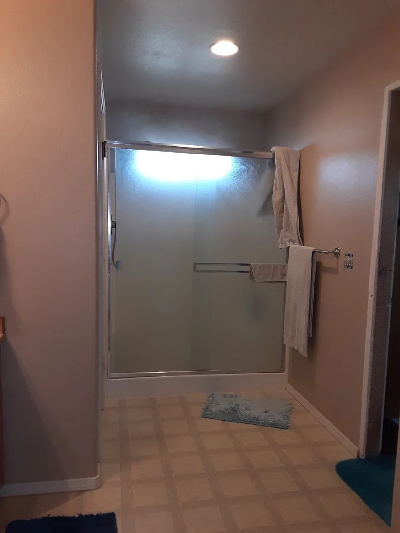 A bathroom with a glass shower door, a towel rack with beige towels, a small blue bath mat on beige tiled floor, and beige walls, with a ceiling light turned on.