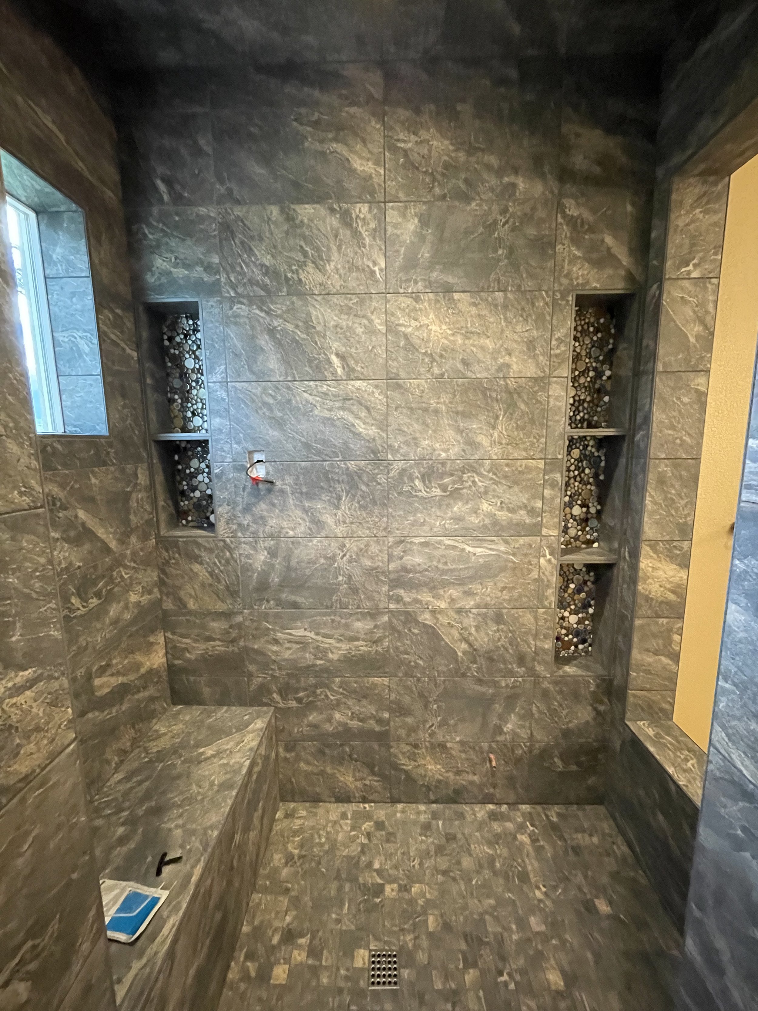 Modern shower with grey marble tiles, built-in shelves filled with small stones, a niche, a bench, and a floor drain.
