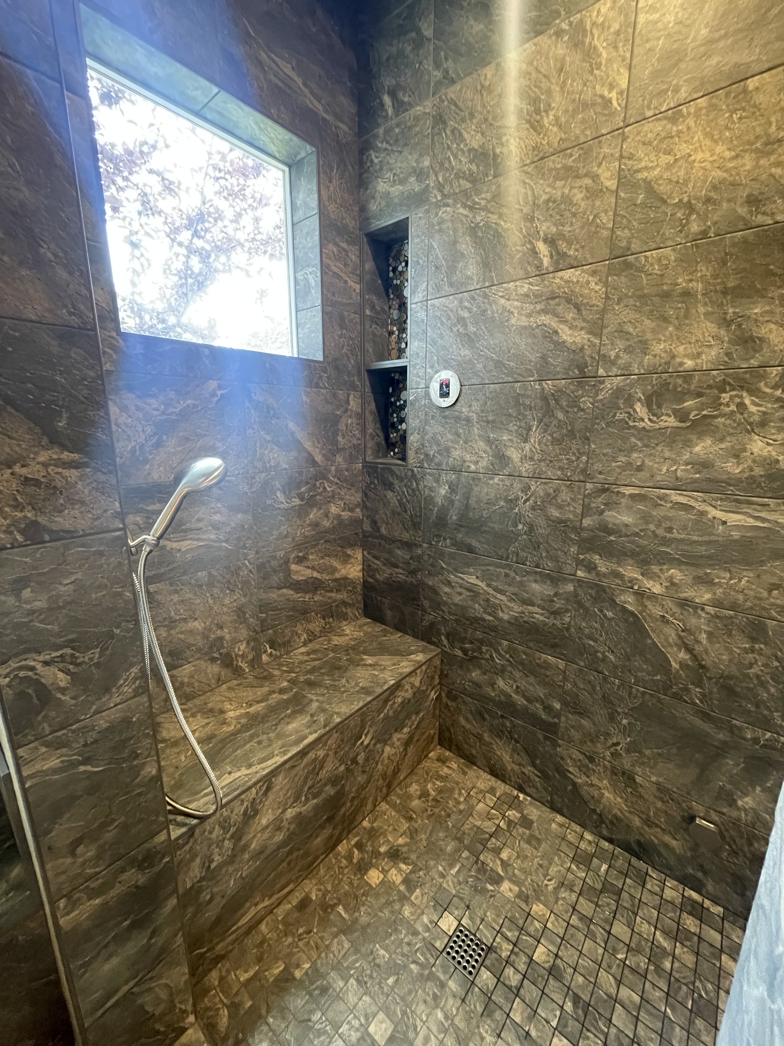 A walk-in shower with dark brown marble tiles, a built-in bench, a narrow window, and a handheld showerhead.