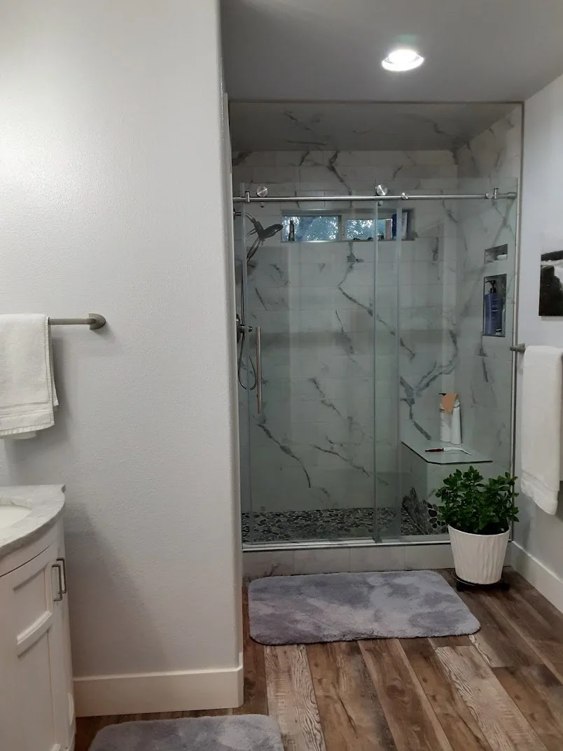 Bathroom with a glass shower enclosure, marble wall tiles, a window, a potted plant, and gray bath mats.