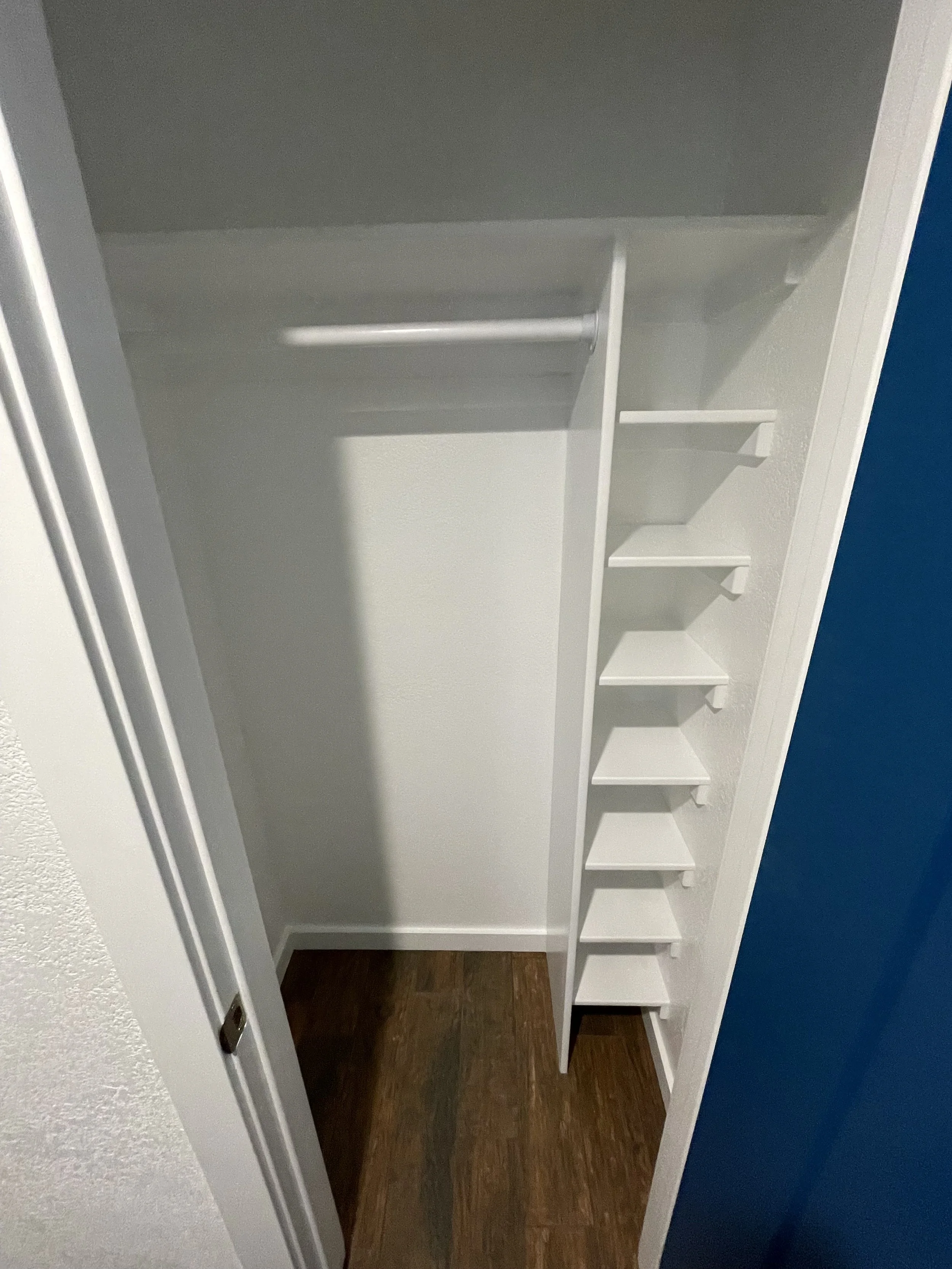 Empty closet with custom white built-in shelves and a hanging rod, dark brown wooden floor, and partially visible blue wall.