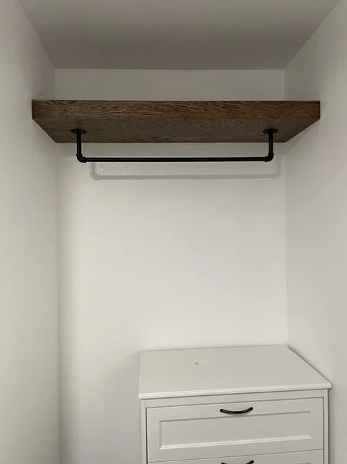 custom built closet with a wooden shelf and an industrial style black clothing rod above a white dresser.
