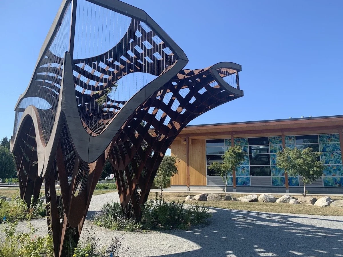 Modern architectural sculpture with curved, open framework made of dark metal near a building with wooden and glass siding, surrounded by small trees and landscaping.