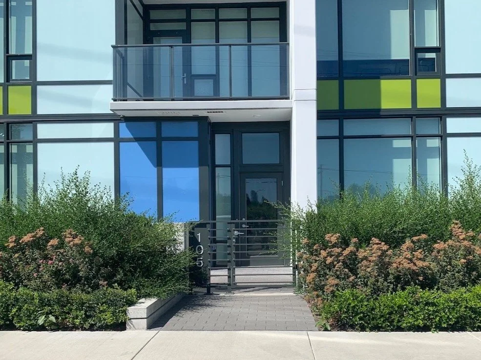 Modern apartment building entrance with glass facade, small balcony, and landscaped bushes along the walkway.