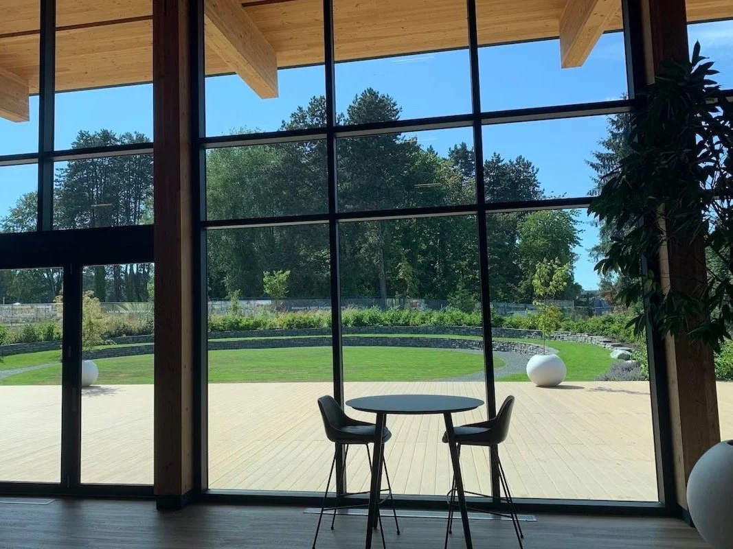 Interior view of a building with large glass windows overlooking a green outdoor area with trees and a grassy lawn. Inside, there is a round table with two black chairs, a wooden ceiling, and a large potted plant.