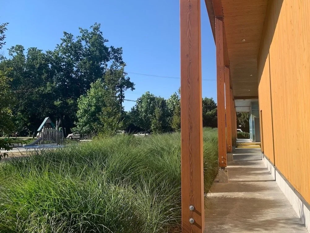 Side view of a building with a wooden exterior, supported by wooden posts, next to a grassy area with tall green grass and a playground in the background, on a sunny day.