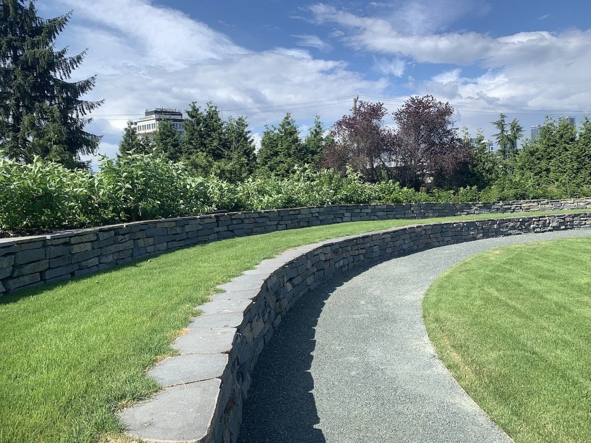 Curved stone pathway running through a well-maintained grassy park with green bushes and trees, under a partly cloudy blue sky.