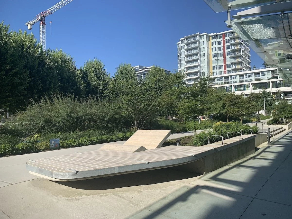 Modern outdoor urban park with a curving concrete and wood seating area and green trees, high-rise residential buildings, and a construction crane in the background, under a clear blue sky.