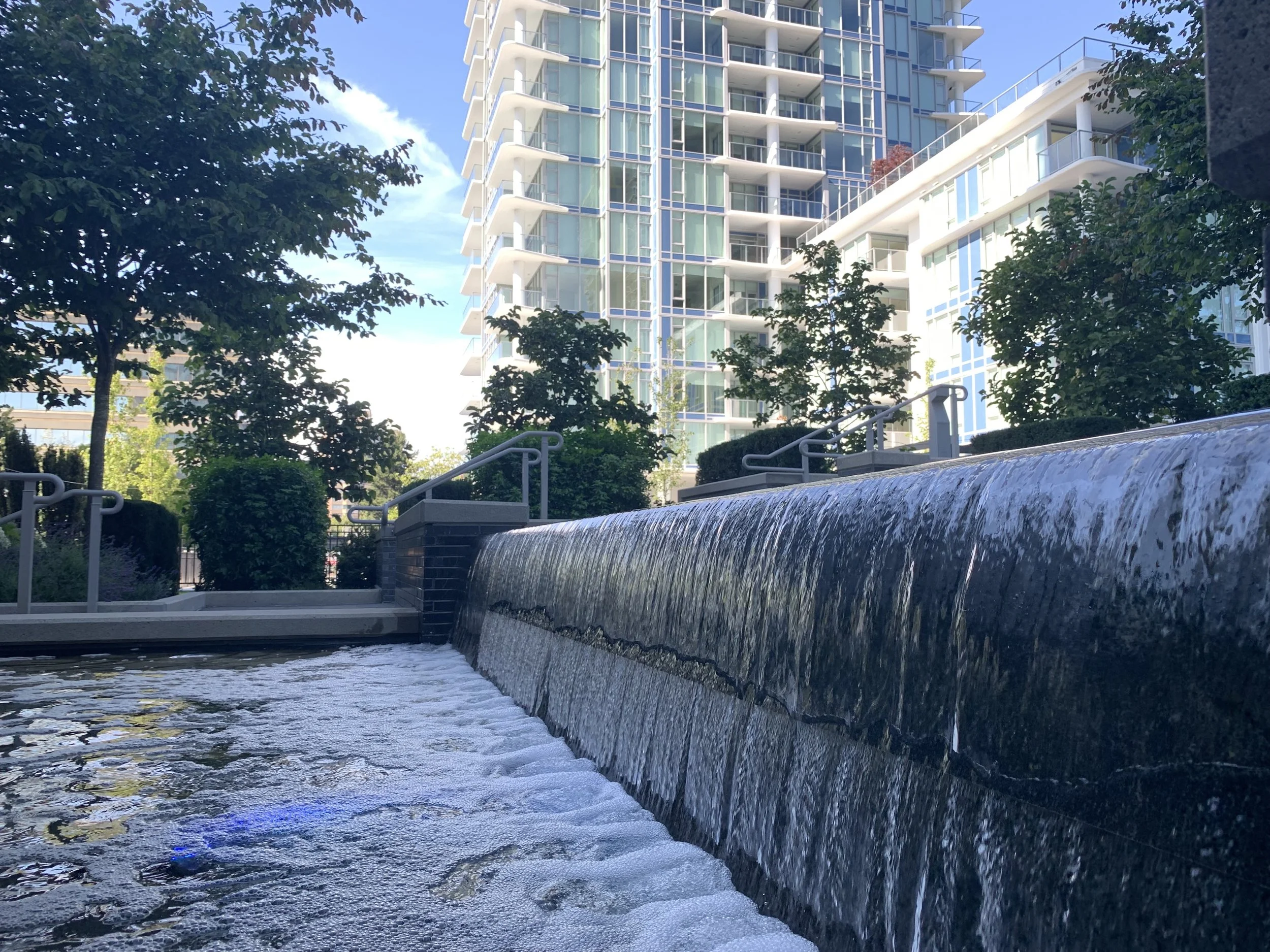 Water feature in an urban park with modern high-rise buildings and trees in the background.