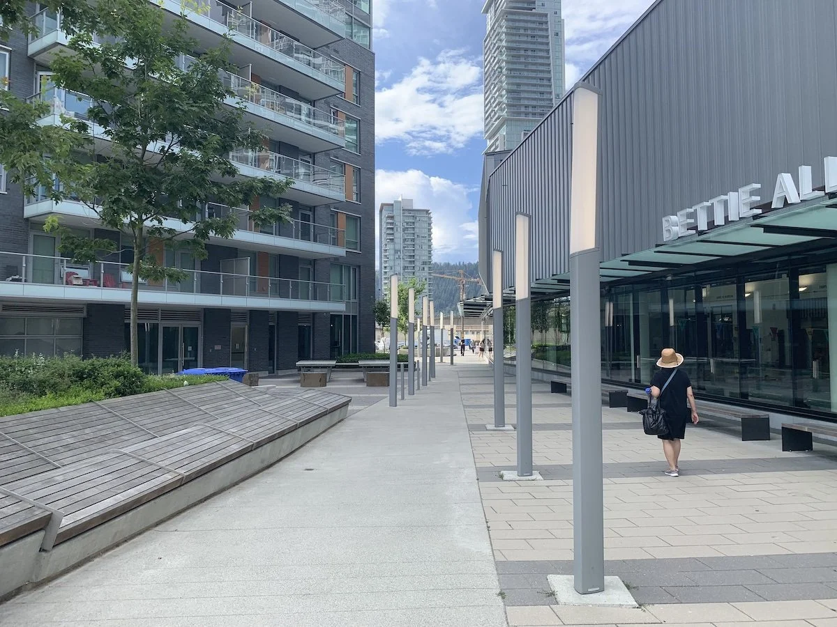 Urban outdoor scene with modern buildings, a sidewalk, benches, trees, and a woman in a black dress and sunhat walking.