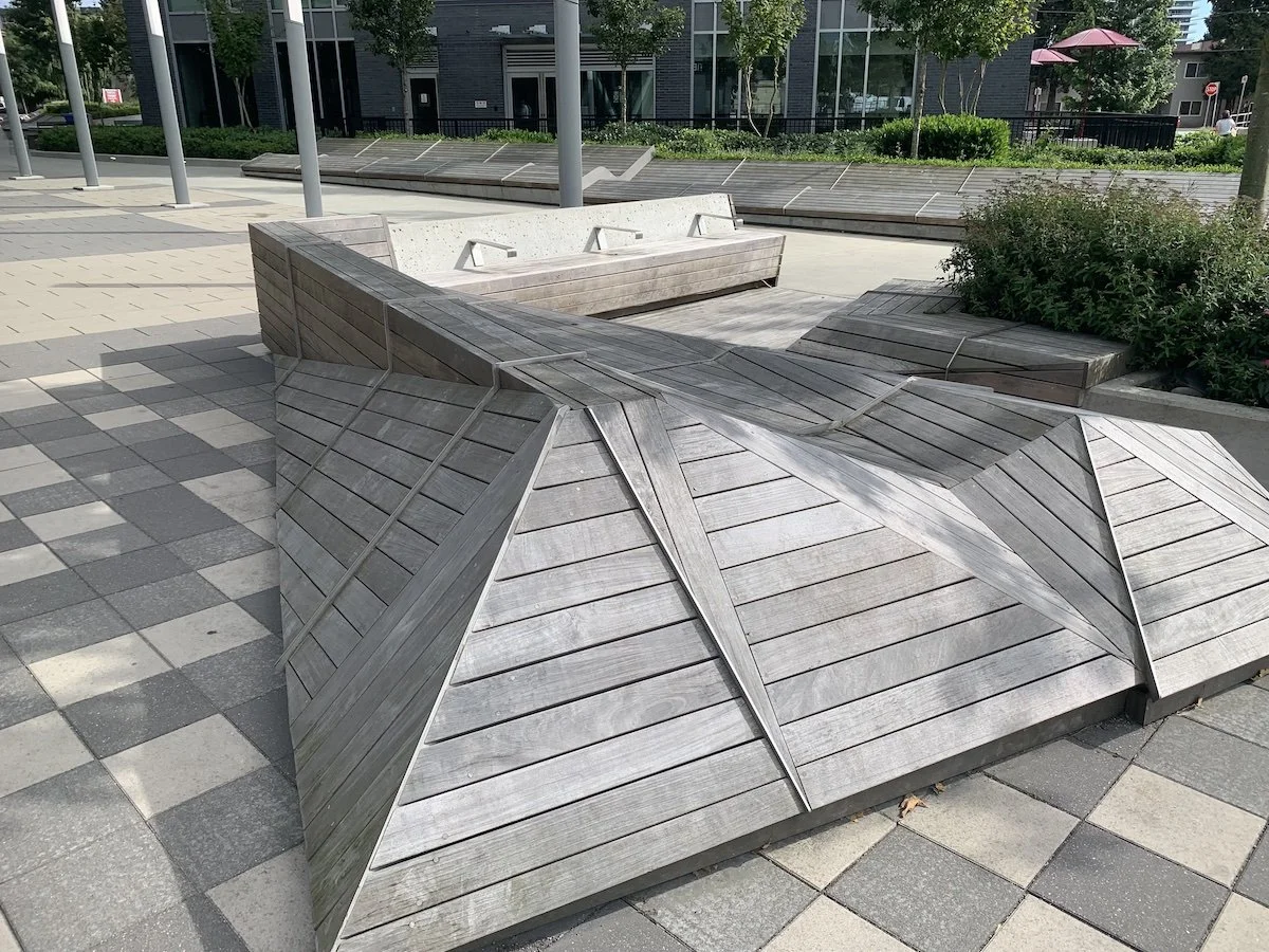 Outdoor public seating area with wooden benches and geometric wooden structures on a paved sidewalk in a modern urban setting.