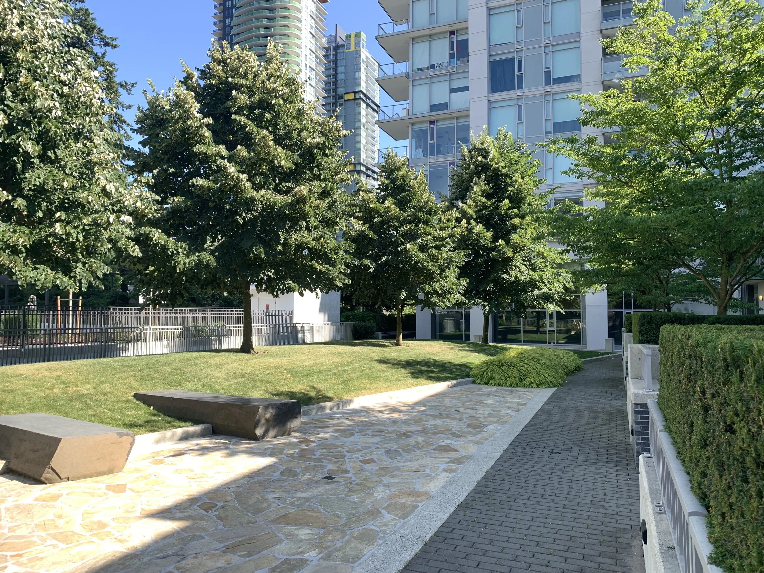 Urban park with green trees, a grassy area, stone and brick pathways, modern glass buildings, and clear blue sky.