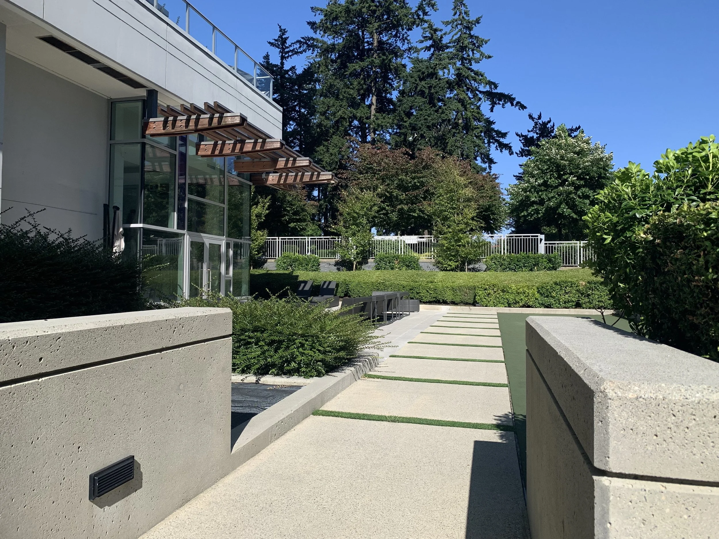 Modern outdoor patio area with concrete walkway, low concrete walls, greenery, and trees with a building featuring glass walls and wooden pergolas in the background.