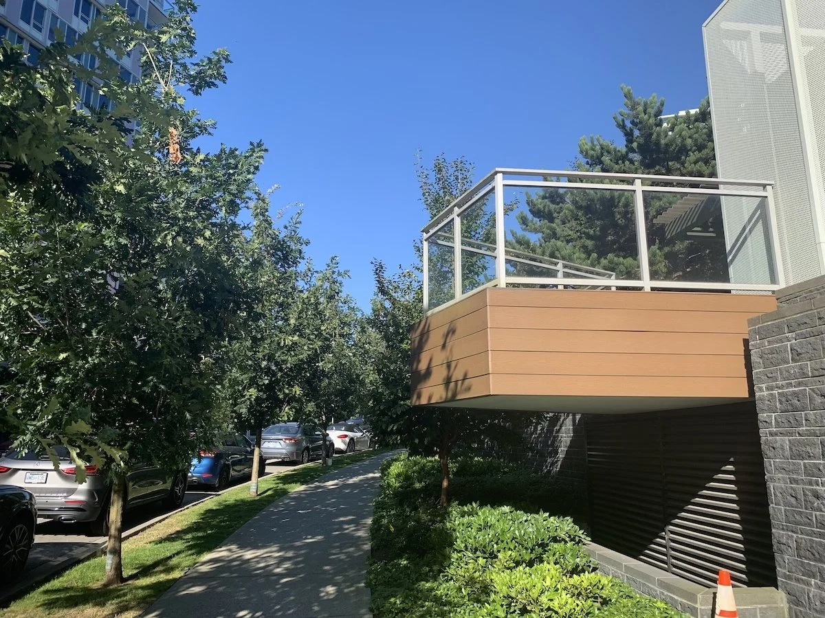 Sidewalk lined with parked cars, trees, and green bushes, alongside an apartment building with a small balcony.