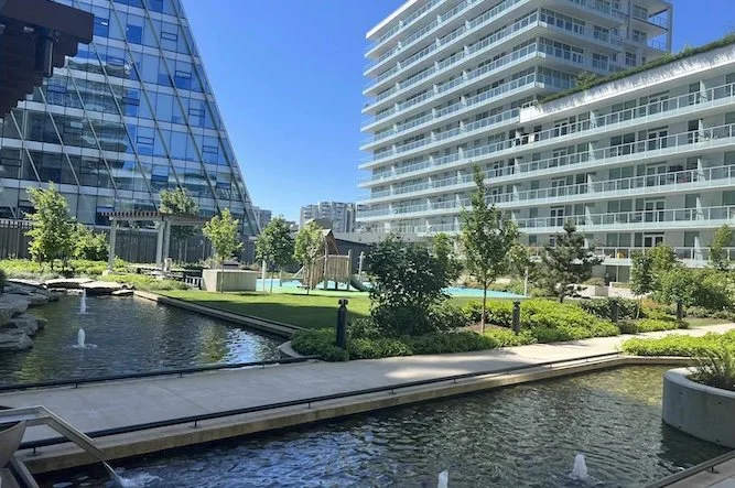 Urban outdoor scene with water features, green landscaping, and modern high-rise buildings under a clear blue sky.