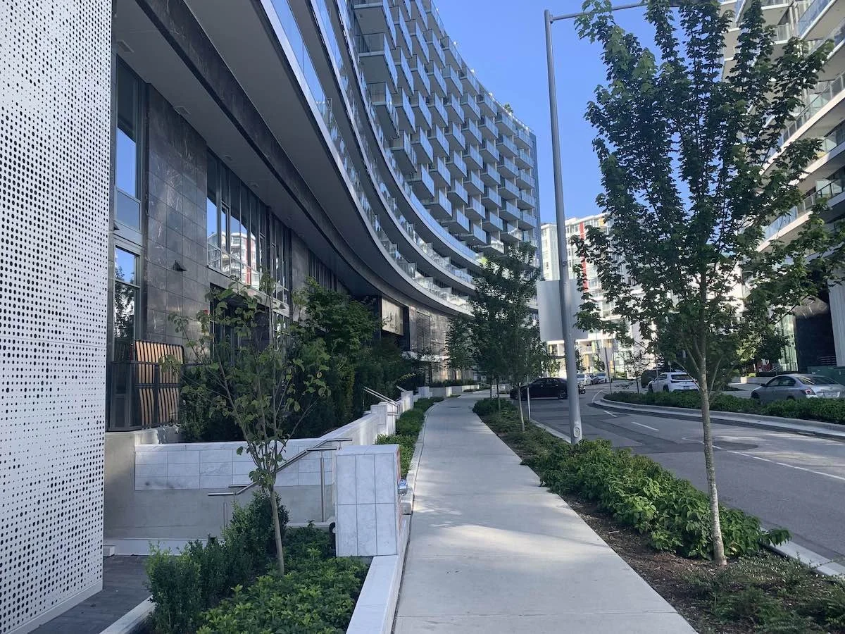 Modern city street with a curved sidewalk, green trees, and tall buildings with reflective windows under a clear blue sky.