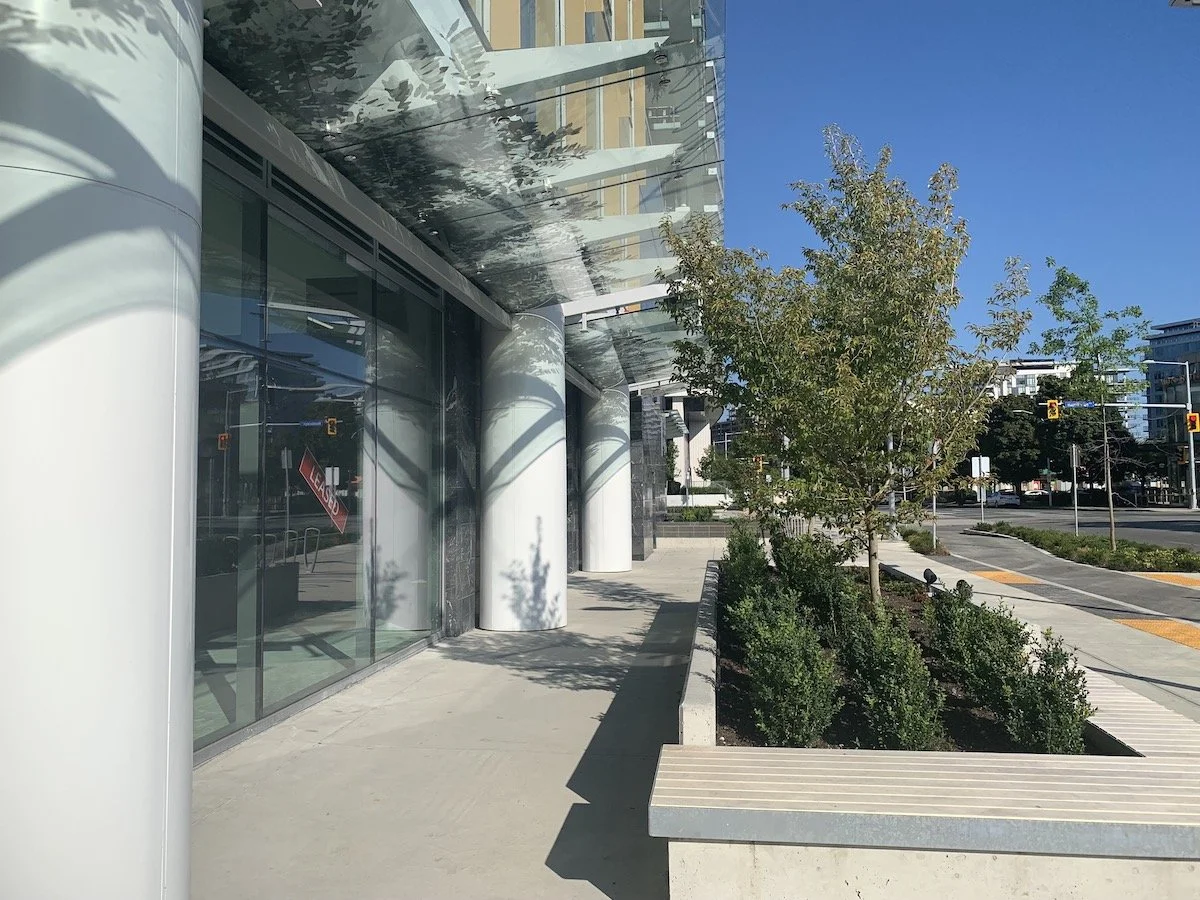 Modern building entrance with large glass doors, white columns, sidewalk, and landscaped trees and bushes, sunny day with a clear blue sky.