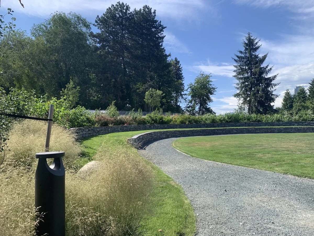 A winding gravel pathway through a well-maintained grassy area, bordered by a stone wall and lush green trees under a partly cloudy sky.
