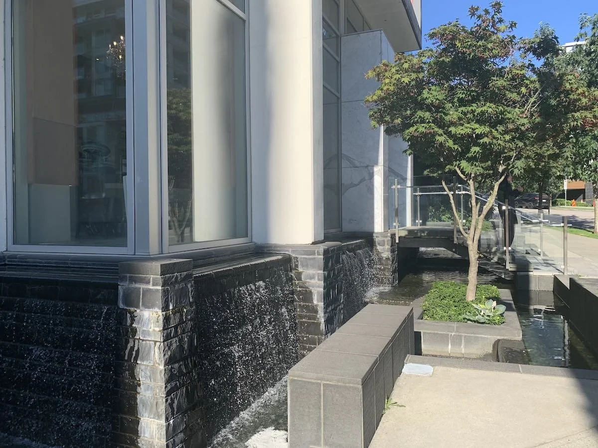 Water feature with cascading water flowing down black tiled walls outside a modern building with large glass windows, a small tree in a square planter, and a sidewalk in the background.