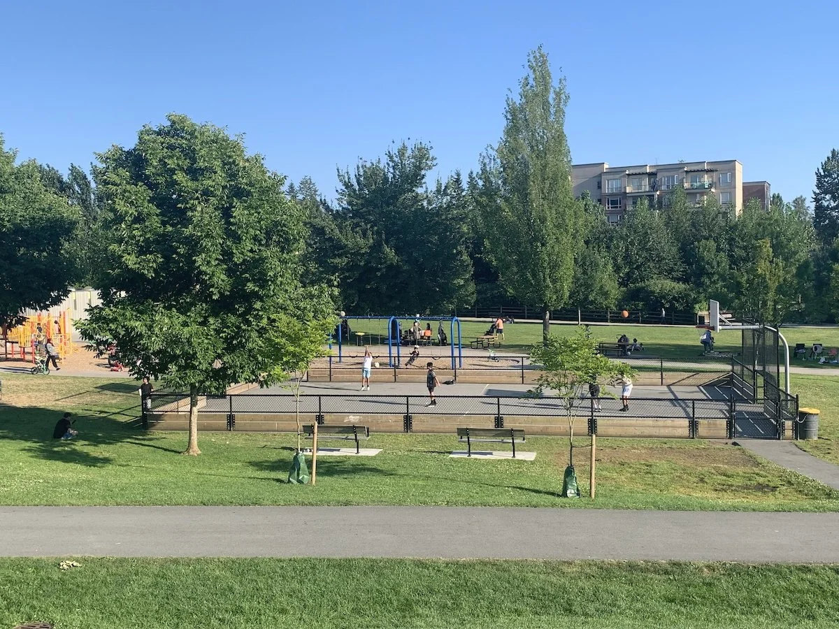 A park with a basketball court and a playground with children playing, surrounded by trees and benches, with a building in the background.