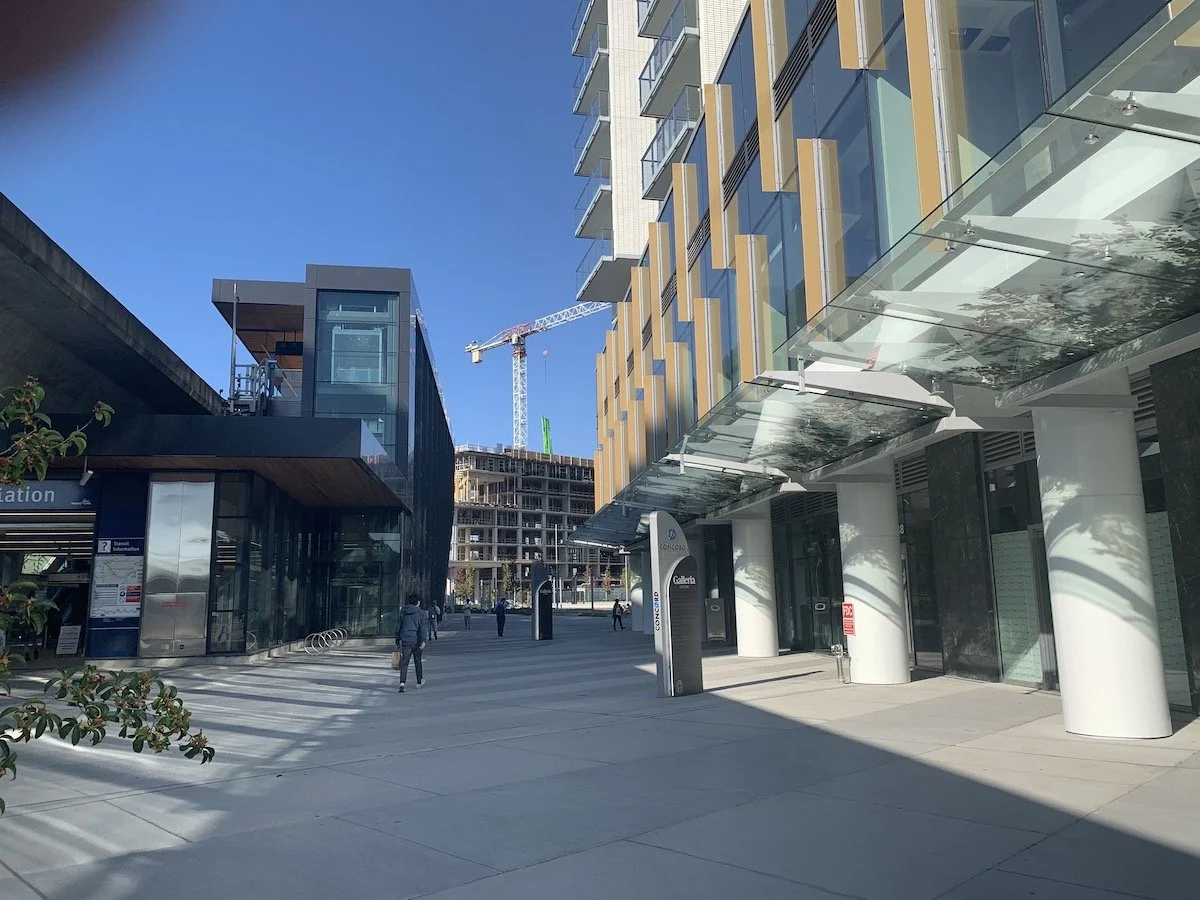 Modern urban area with glass buildings, a construction crane in the background, and a few pedestrians walking on a wide sidewalk under clear blue skies.