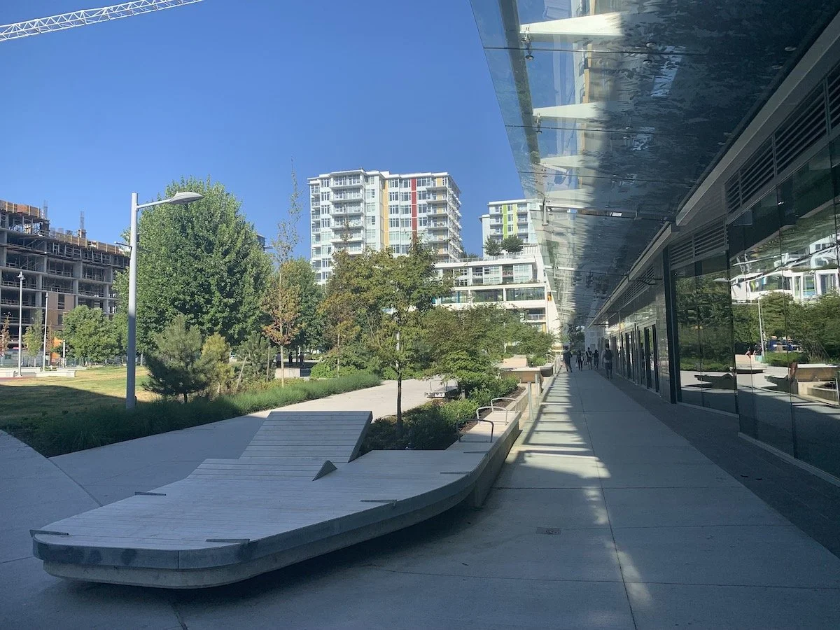 Modern urban park with trees, benches, and a glass building reflecting the surroundings on a bright sunny day.