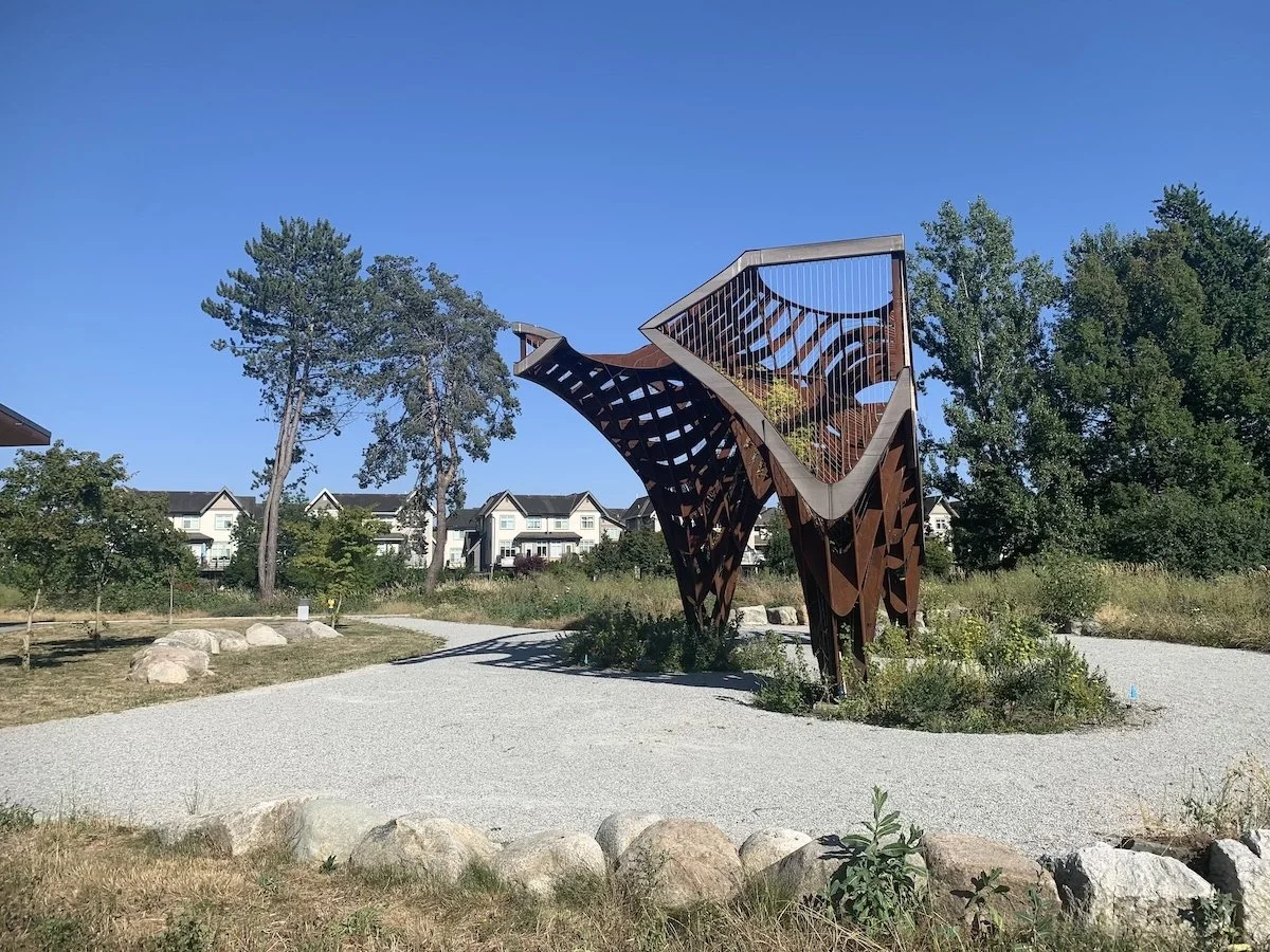 A modern, abstract sculpture made of metal with geometric cutouts, located in a landscaped park with trees and residential buildings in the background.