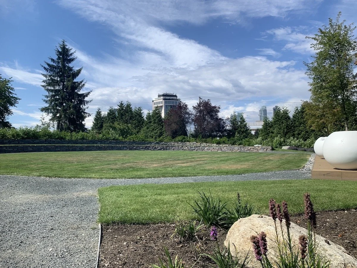 A landscaped park with trees, a grassy area, a stone wall, and large white planters, with city skyscrapers visible in the background under a partly cloudy sky.