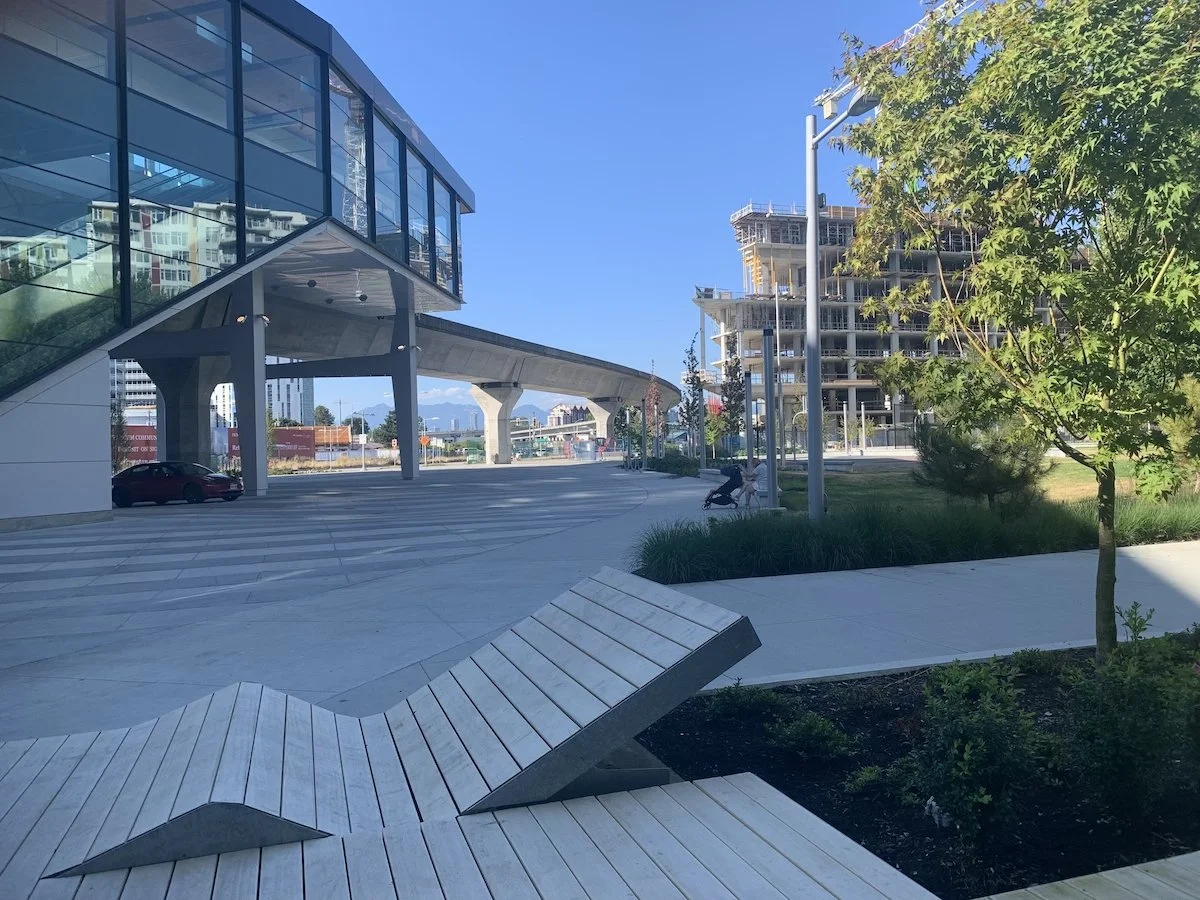 Modern urban scene with a glass building on the left, a construction site with a multi-story building on the right, a curved walkway underneath, a bench in the foreground, and a person pushing a stroller on the sidewalk. Clear blue sky.