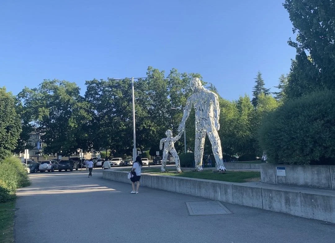 A large outdoor sculpture of a man holding the hand of a smaller child is depicted, with trees and a parking lot in the background.