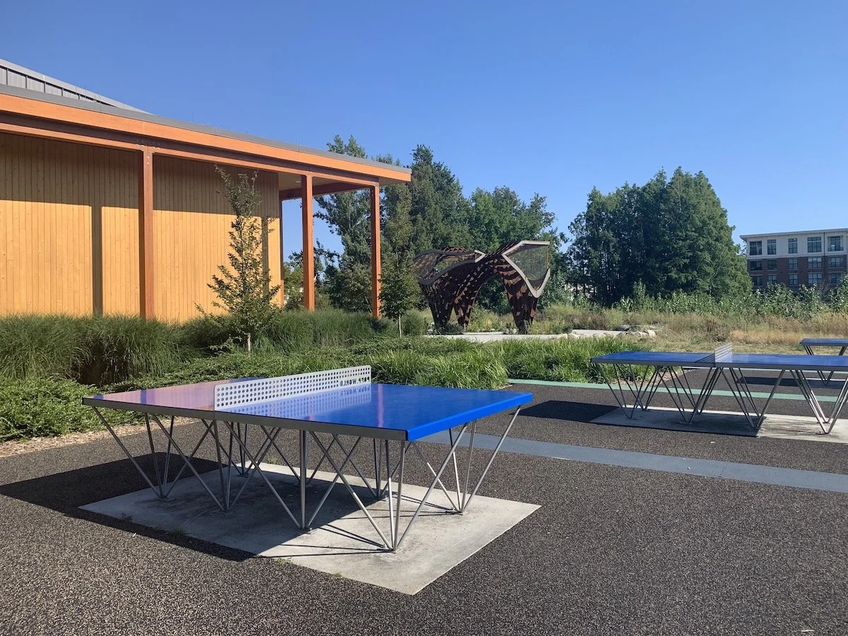 Outdoor scene with two blue ping pong tables on a paved court, green grass and trees, a modern wooden building with orange accents, and a metal sculpture in the background under a clear blue sky.
