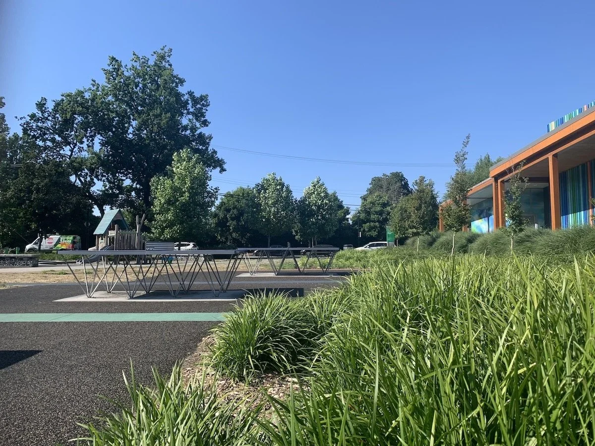 Empty outdoor parking lot with metal bike racks, green grass, trees, a clear blue sky, and a modern building with colorful panels on the right.