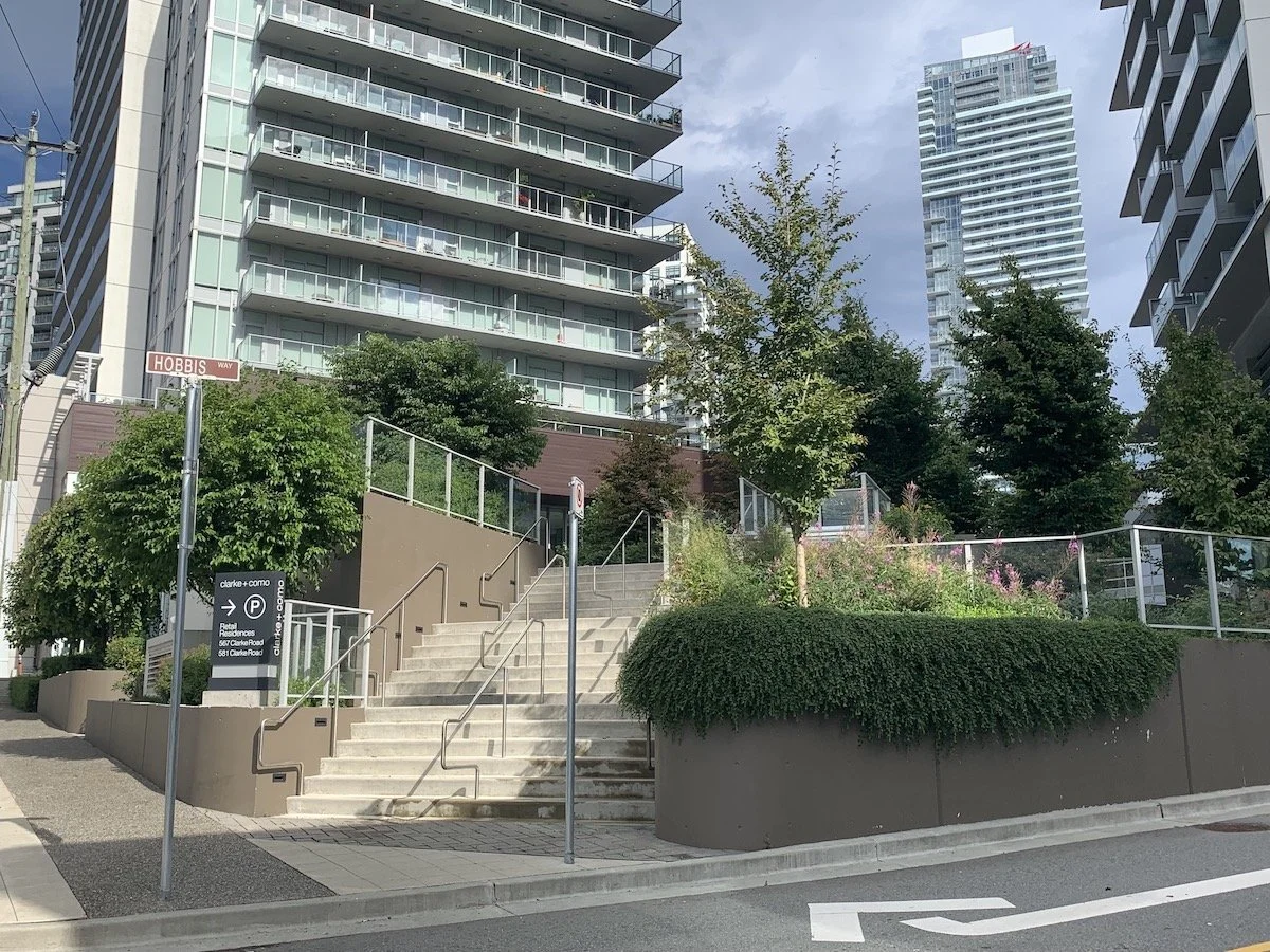 City street corner with stairway leading to a building entrance, surrounded by trees and modern high-rise buildings in background, and directional signs including one for parking.