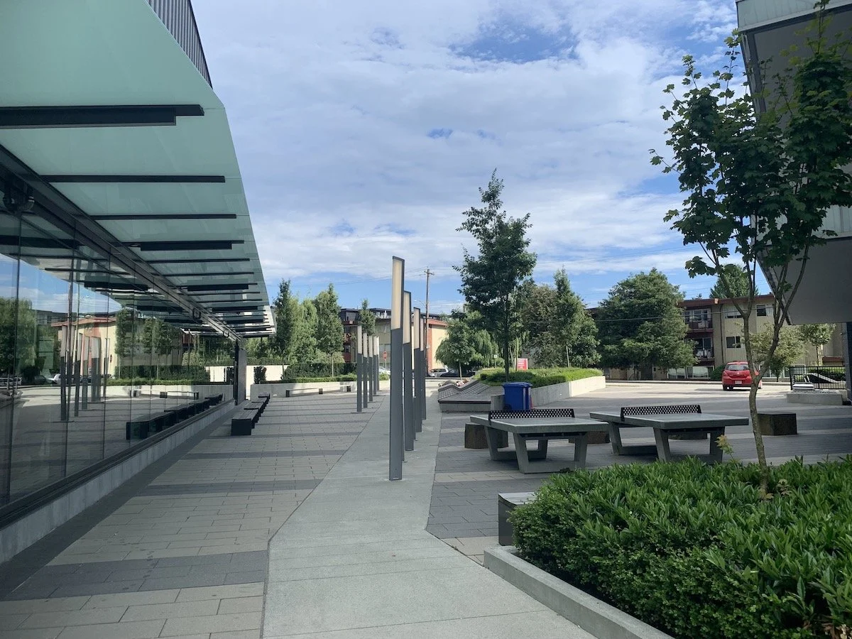 Empty outdoor plaza area with benches, trees, a building with reflective glass windows, and a cloudy sky.