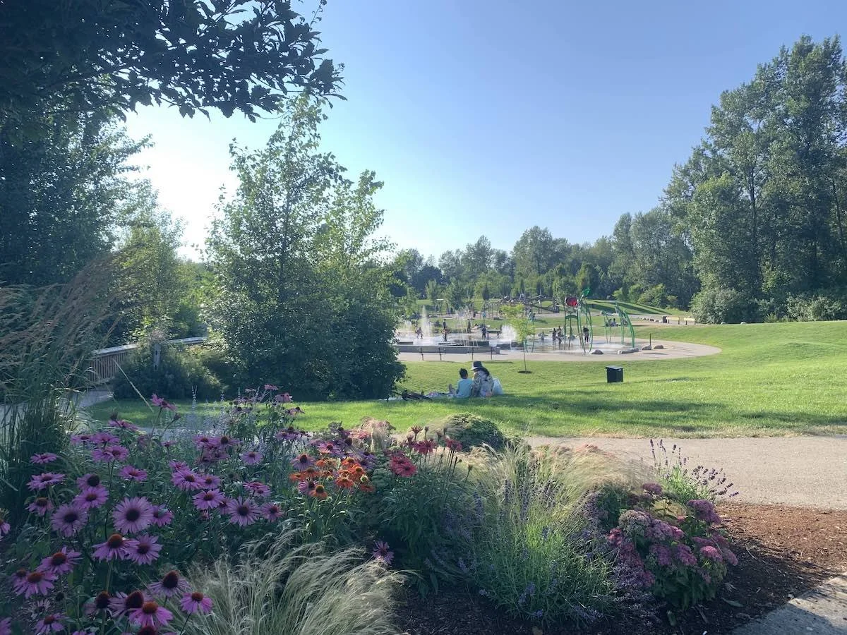 A park with a water spray area for children, green grass, trees, and a flower bed in the foreground. People are sitting on the grass near the fountain, and others are playing in the water feature on a sunny day.