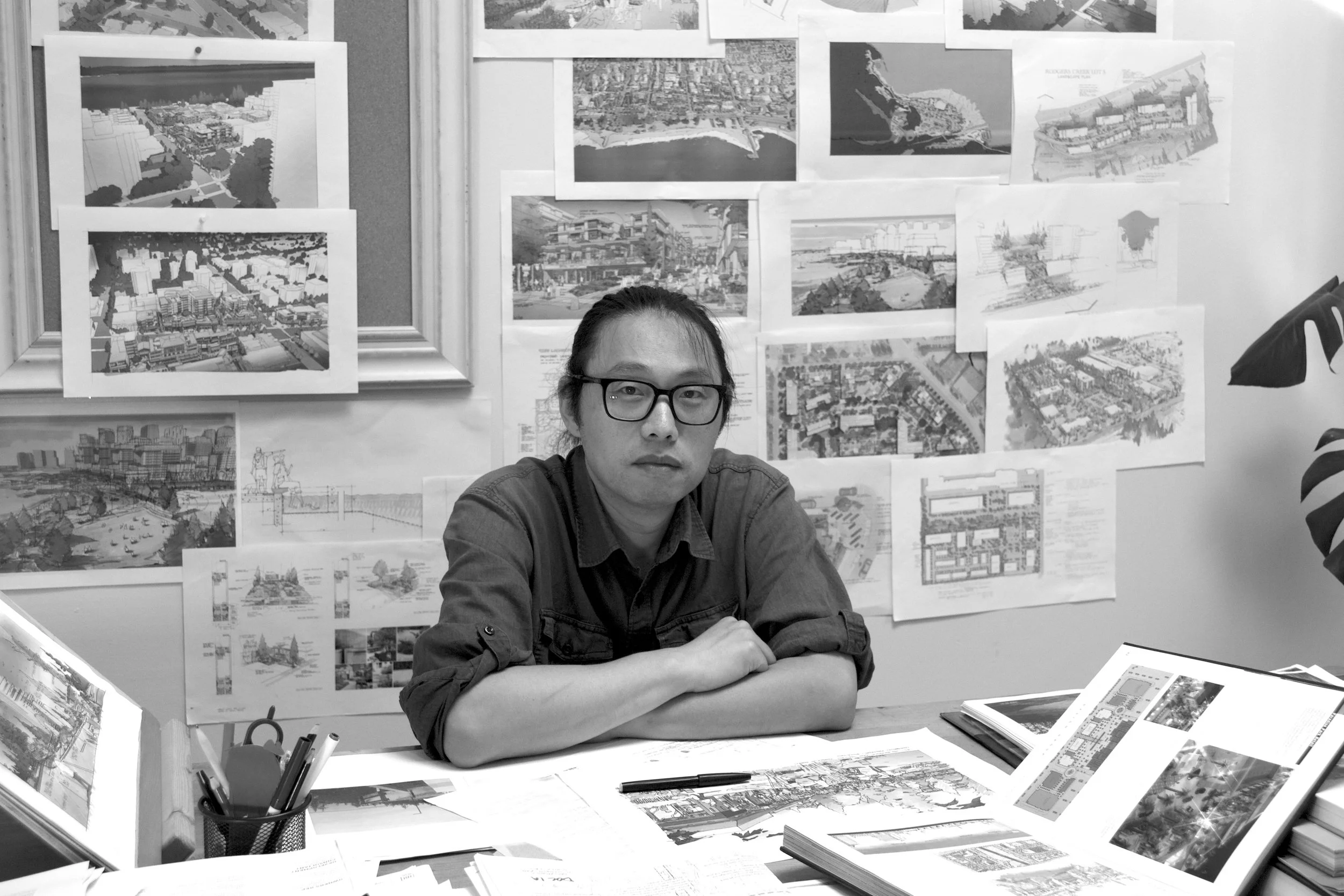A person with glasses and long hair tied back sitting at a desk in front of a wall covered with architectural drawings, plans, and renderings, with open books and papers on the desk.