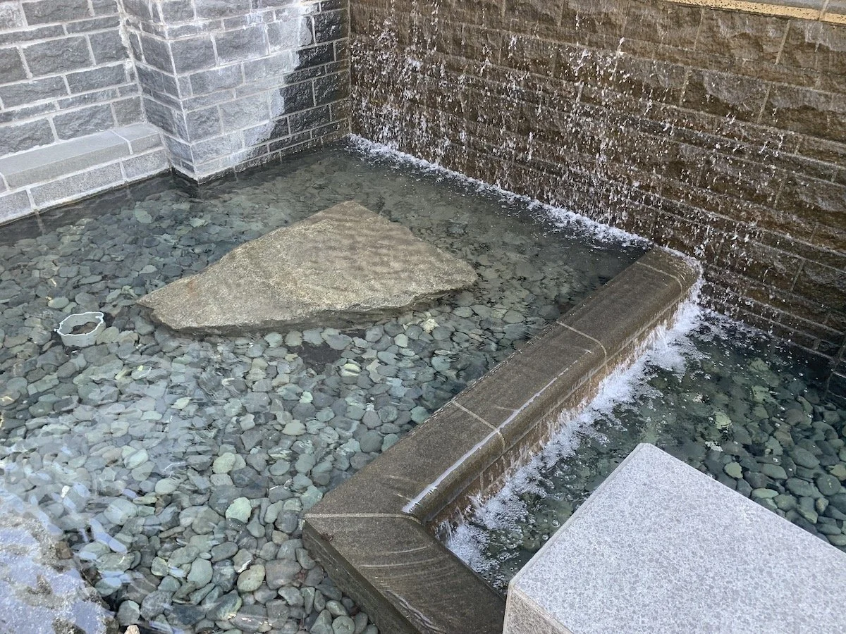A small outdoor water feature with a stone border, a large flat rock in the shallow water, and a wall with water flowing over it.