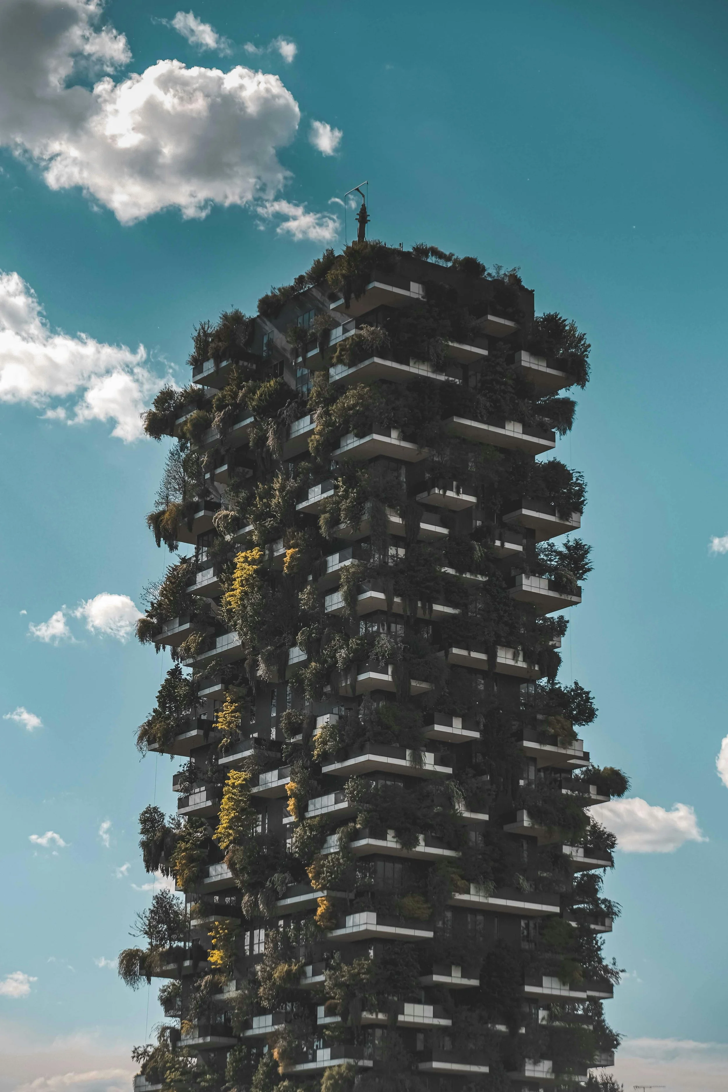 A tall building with multiple floors covered in lush green plants and trees on balconies, against a blue sky with a few scattered clouds.