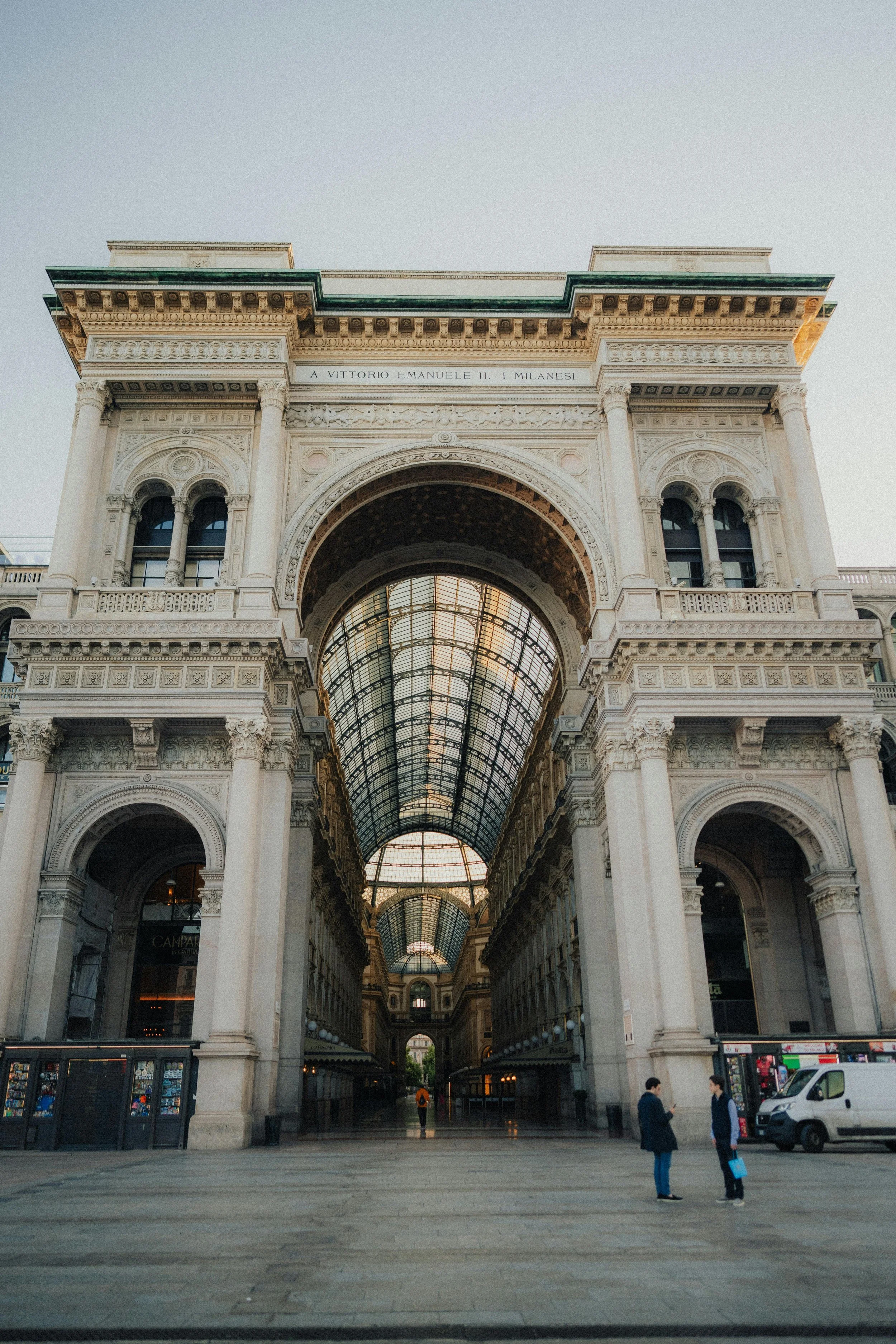 Historic building with an arched glass roof in Milan, Italy, with two men in conversation in the foreground.