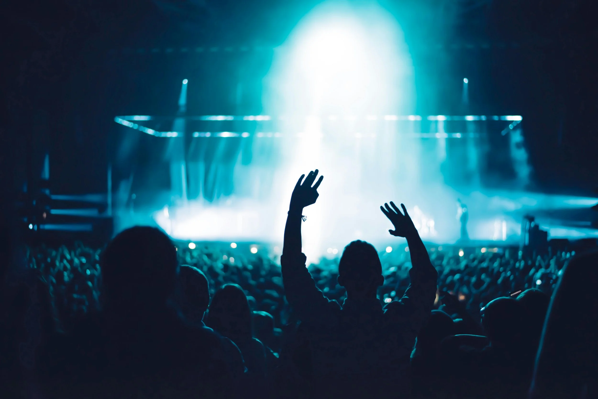 A concert crowd with a person raising their hands in front of a brightly lit stage with blue lighting.