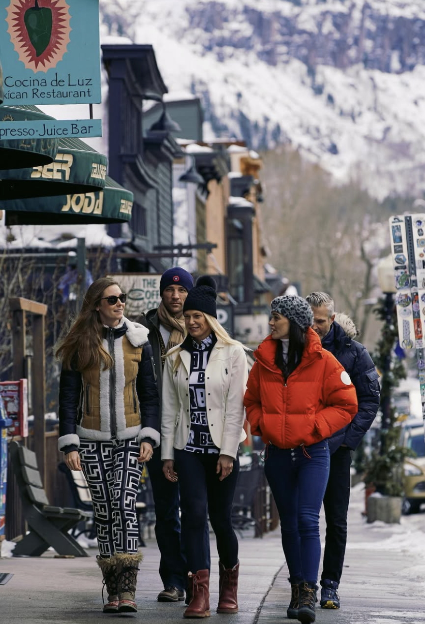 Five people walking on a snowy street in a mountain town, dressed in winter clothing, with shops and snow-covered mountains in the background.