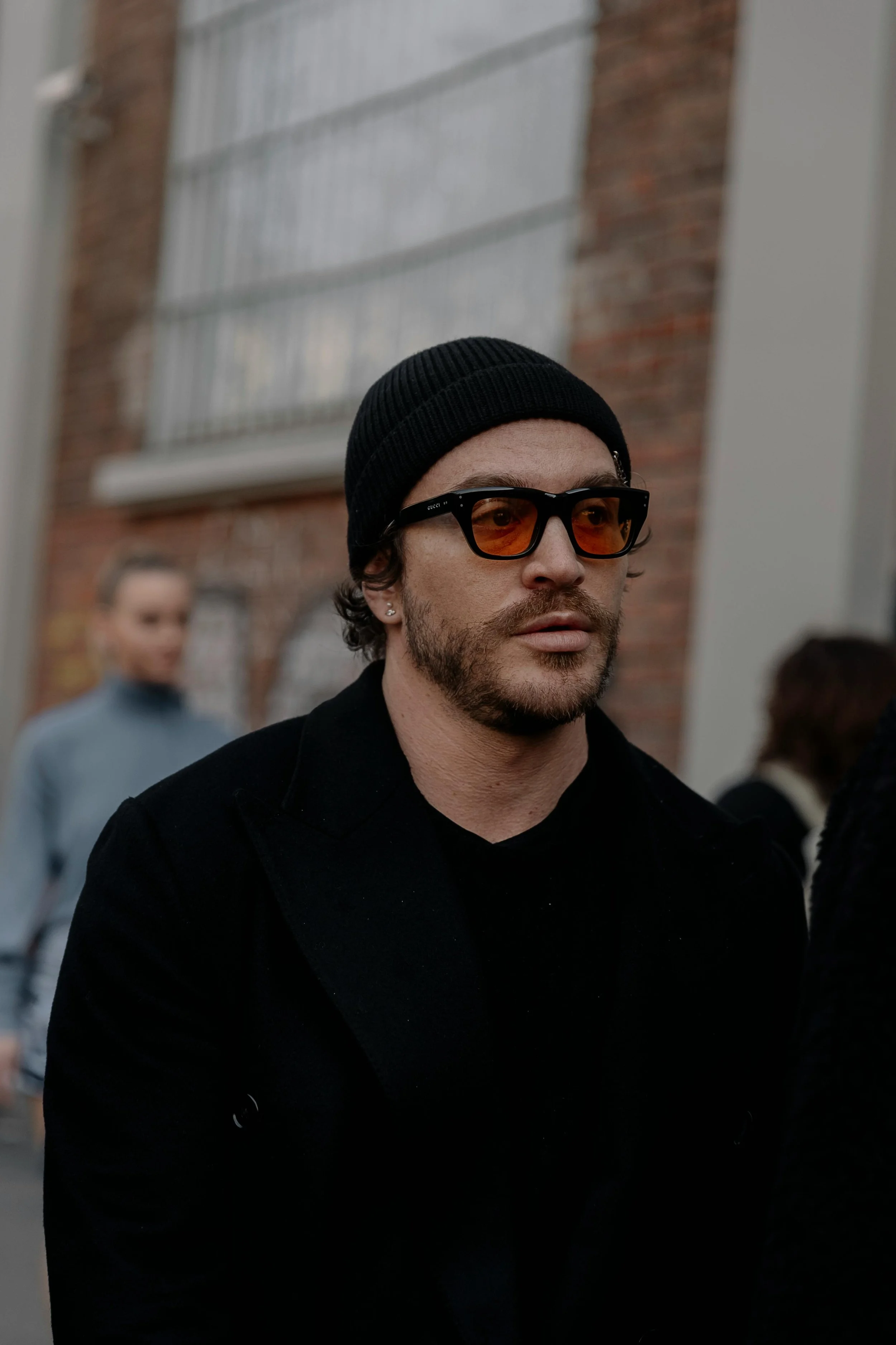 A man with a beard and curly hair wearing a black beanie, orange-tinted sunglasses, and a black coat, walking outdoors in front of a brick building with a woman blurred in the background.