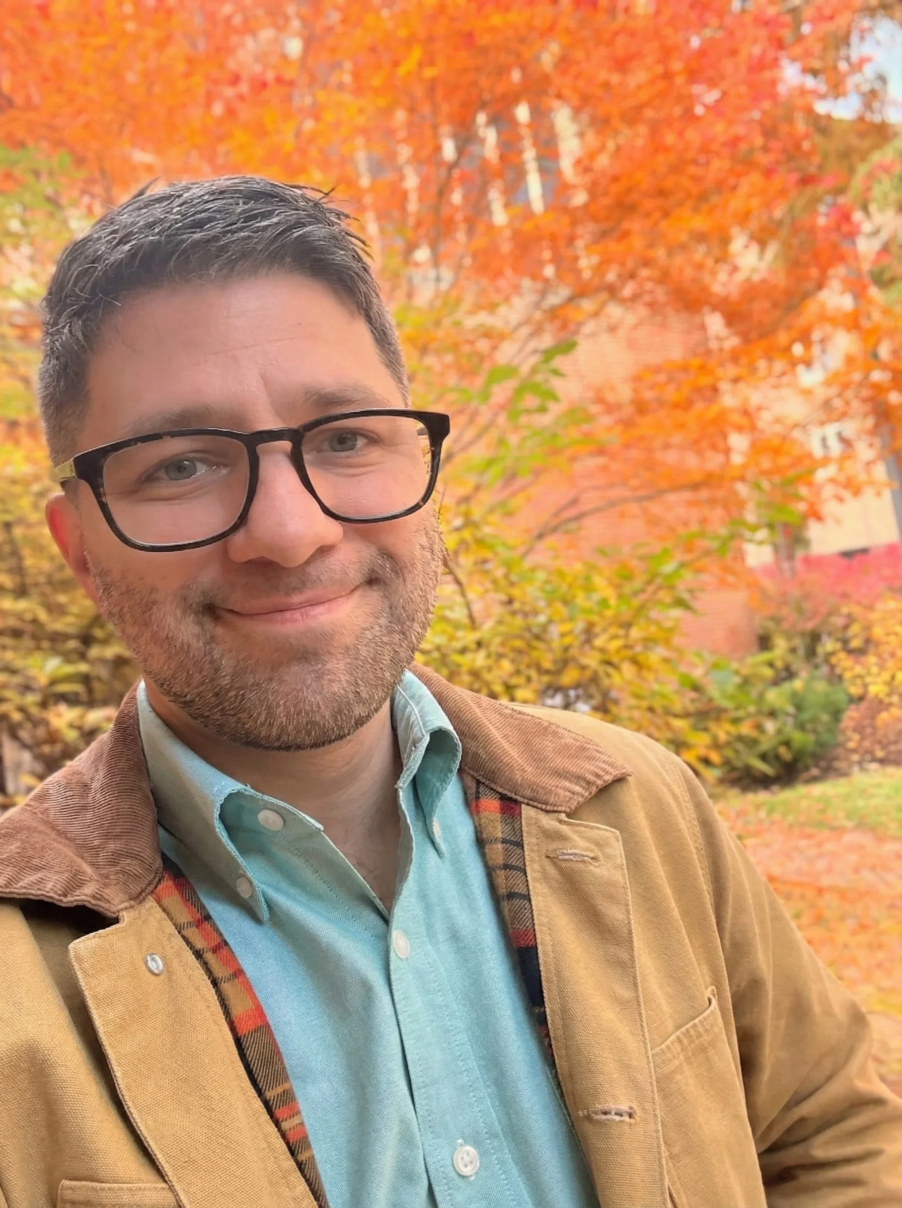 A man with glasses and a beard taking a selfie outdoors during autumn, with trees having vibrant orange and yellow leaves in the background.