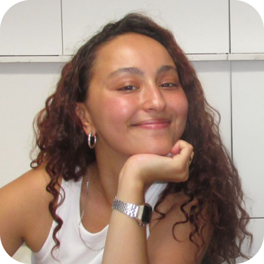 Jeune femme souriante avec des cheveux bouclés, portant un bracelet et des boucles d'oreilles, assise devant un mur blanc.