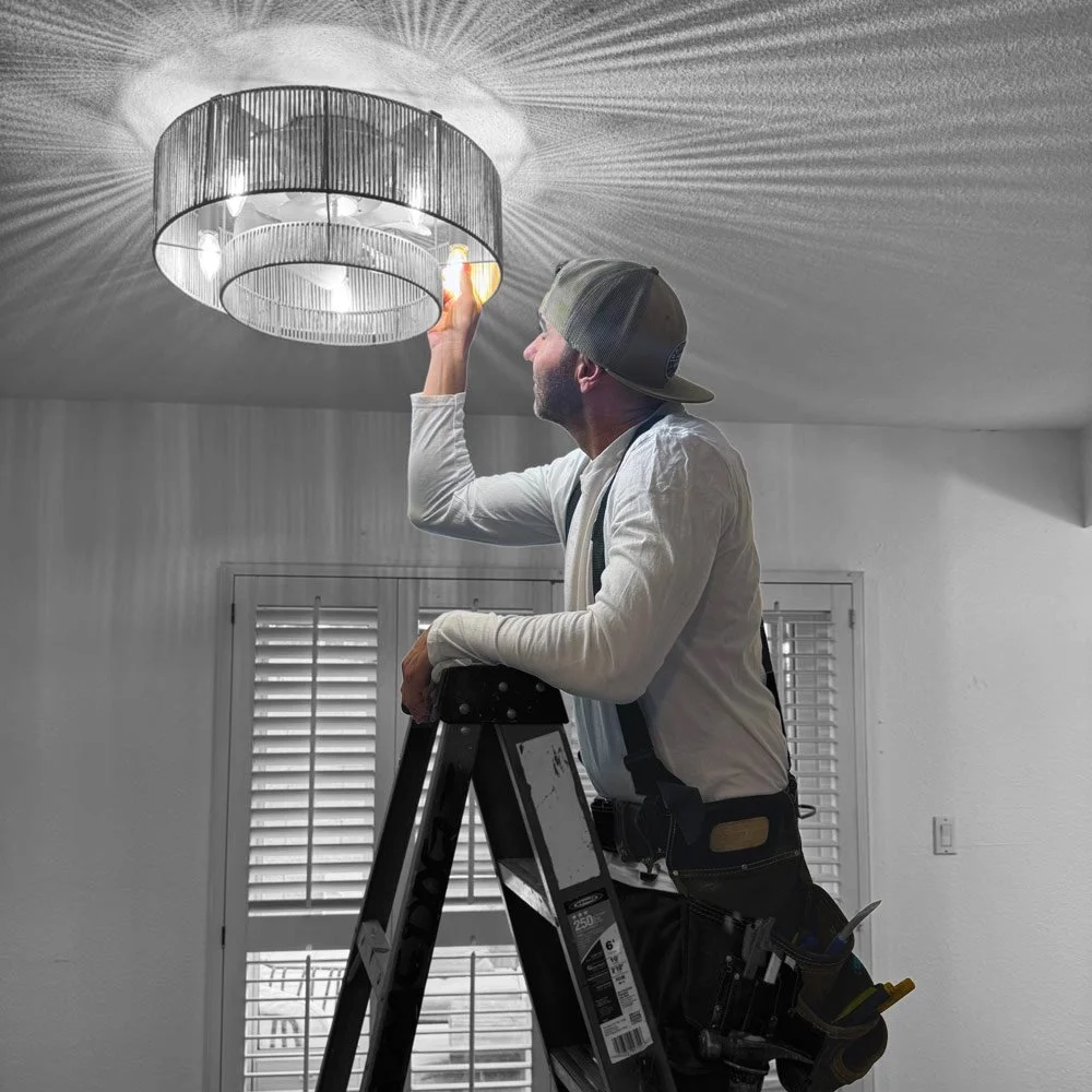 A man in a baseball cap, white shirt, and tool belt is standing on a ladder adjusting a chandelier that is casting intricate shadows on the ceiling.