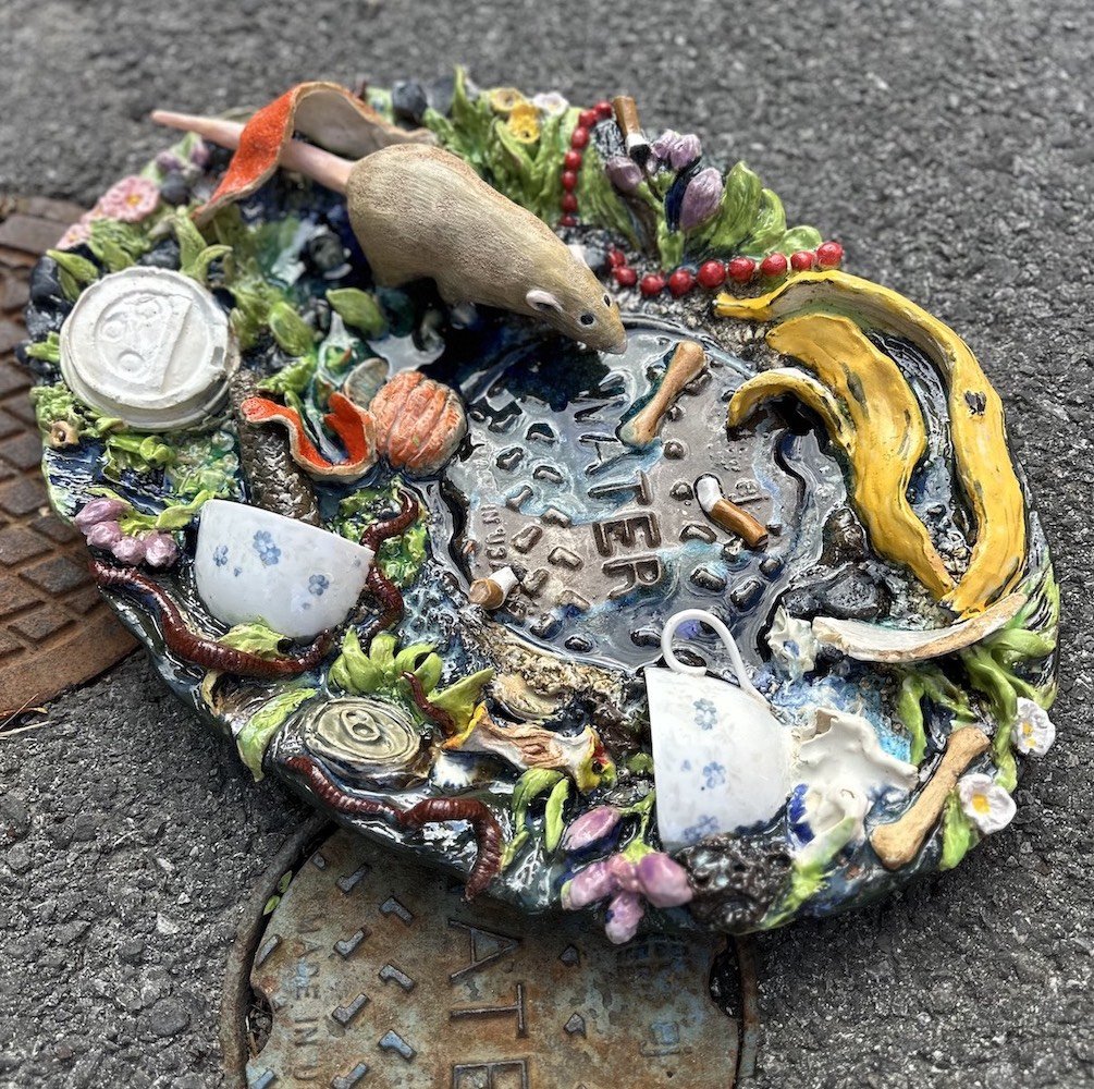 Colorful ceramic sculpture of a mouse near a small pool with the word 'NEVER' written inside, surrounded by flowers, vegetables, and other small objects on a street surface.